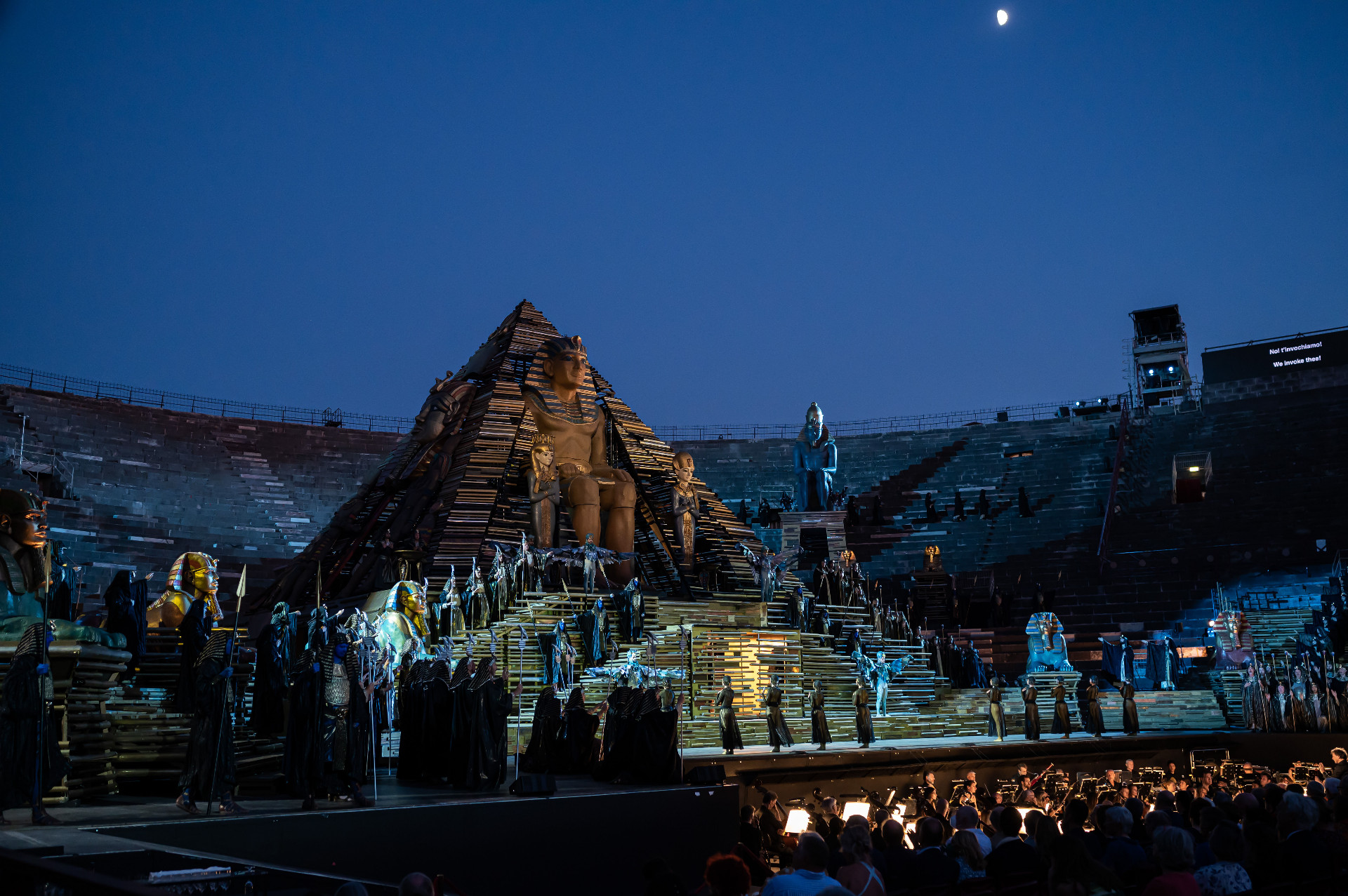 Radamès e Aida, in ginocchio l’uno accanto all’altra, sono all’interno di una struttura in ferro a forma di piramide, che ricorda quella del Louvre. Sono entrambi vestiti di bianco e tendono le mani in avanti. Aida ha la fronte truccata di rosso chiaro e di lilla, decorata con dei glitter colorati, mentre quella di Radamès, al centro, è decorata con una piccola riga nera.