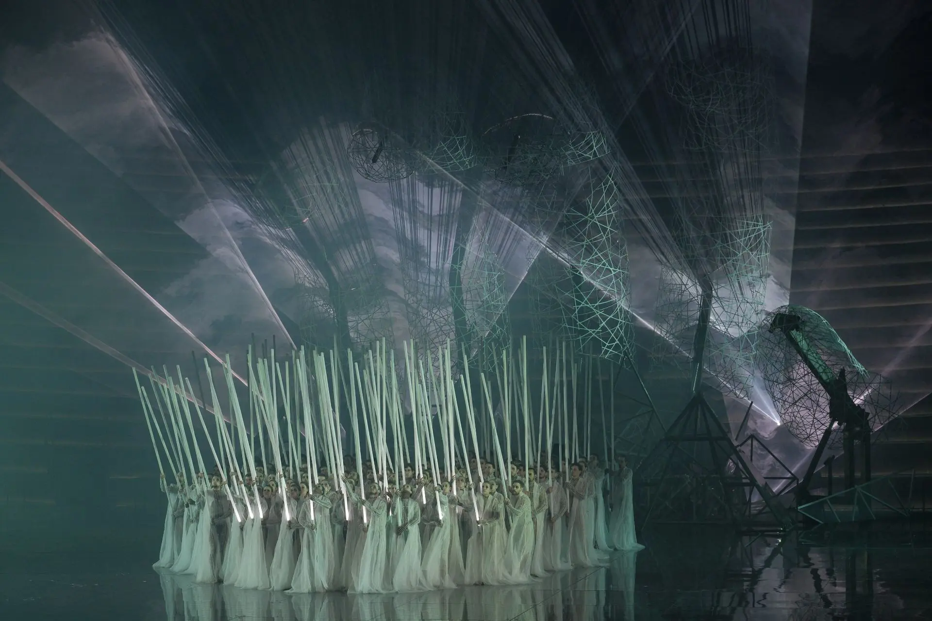 Il palcoscenico è ancora dominato dalla grande mano, illuminata da fasci di luce bianca e circondata da dense nuvole di fumo. I danzatori e le danzatrici sono in piedi, l’uno accanto all’altra, e occupano il centro del palcoscenico. Sopra alla tuta indossano ora una lunga tunica di tulle bianco. In mano stringono un lungo bastone illuminato di bianco, simile ad una spada laser, che rivolgono verso l’alto.