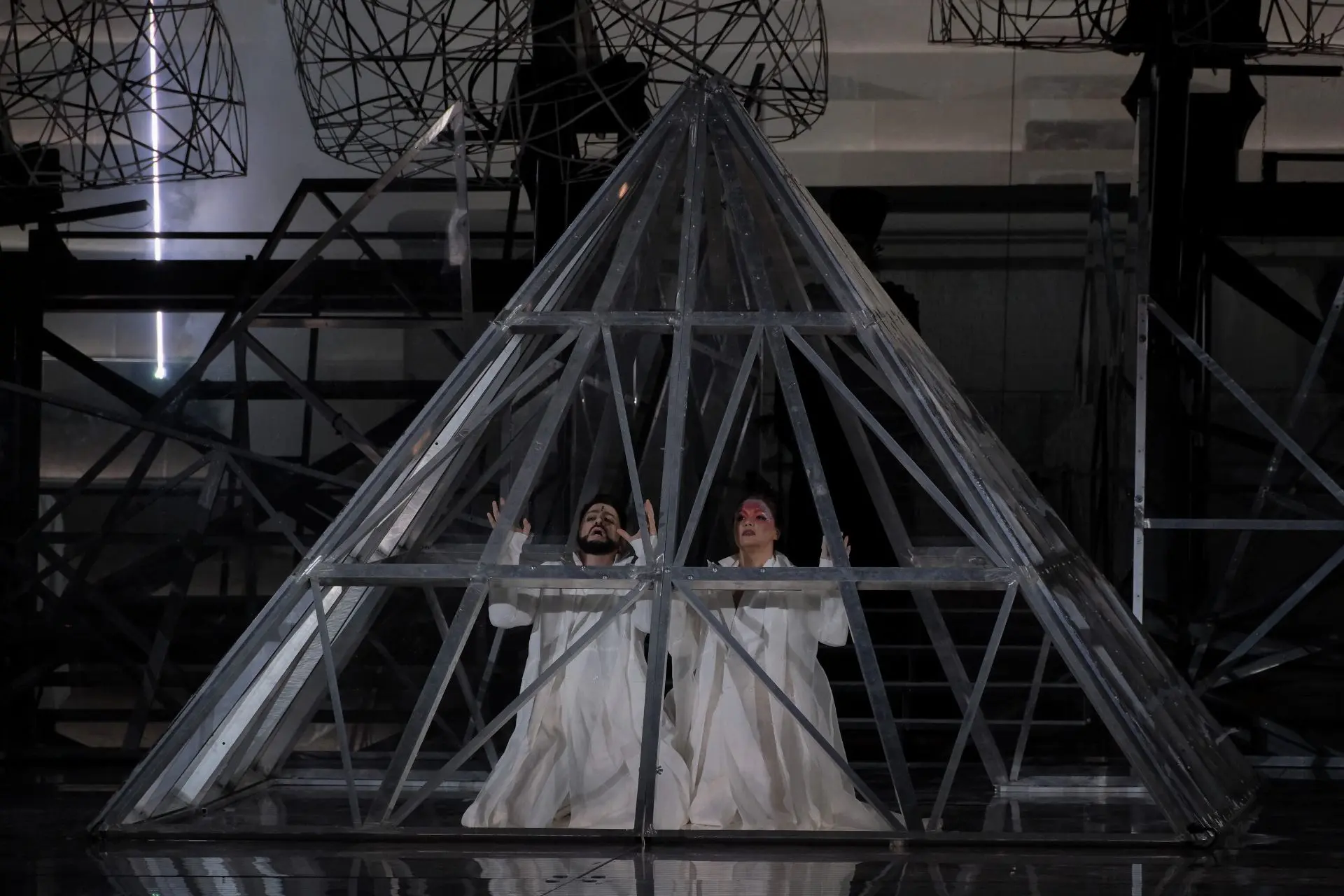 Radamès e Aida, in ginocchio l’uno accanto all’altra, sono all’interno di una struttura in ferro a forma di piramide, che ricorda quella del Louvre. Sono entrambi vestiti di bianco e tendono le mani in avanti. Aida ha la fronte truccata di rosso chiaro e di lilla, decorata con dei glitter colorati, mentre quella di Radamès, al centro, è decorata con una piccola riga nera.
