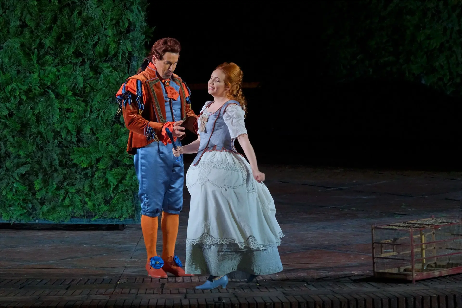 Figaro e Rosina sono l’uno accanto all’altra, al centro della scena, di fronte al labirinto. Figaro indossa un completo di raso, di colore azzurro vivo, composto da giacca corta e abbottonata e pantaloni lunghi fino alle ginocchia, con sotto delle calze arancioni. Sopra alla giacca indossa una casacca di velluto, di colore arancione, tenuta aperta. La casacca è più corta e dritta nella parte frontale, più lunga e plissettata sul retro. Le spalline sono rinforzate e decorate con degli inserti azzurri e arancioni, mentre i polsini e il colletto sono decorati con dei volant, di colore arancione acceso. Ai piedi, scarpe arancioni di forma squadrata, con il tacco basso, decorate con un grande fiore blu all’altezza del collo del piede. Indossa una parrucca castana, con i capelli raccolti sul retro in una coda legata con un nastro. Rosina indossa un ampio abito di cotone leggero, lungo fino alle caviglie. La parte superiore è composta da un corpetto rinforzato, di colore celeste scuro, con i bordi rosso scuro; le maniche sono corte, di colore celeste chiaro. La gonna, di colore celeste chiaro, è ampia, a strati; quello superiore è decorato da merletti. Ai piedi, scarpe celeste scuro di forma squadrata, con il tacco basso. I capelli sono castano chiaro, quasi arancioni. Sono raccolti sulla nuca e tenuti sciolti lungo le spalle. Stringe in mano un ventaglio chiuso, di colore blu.