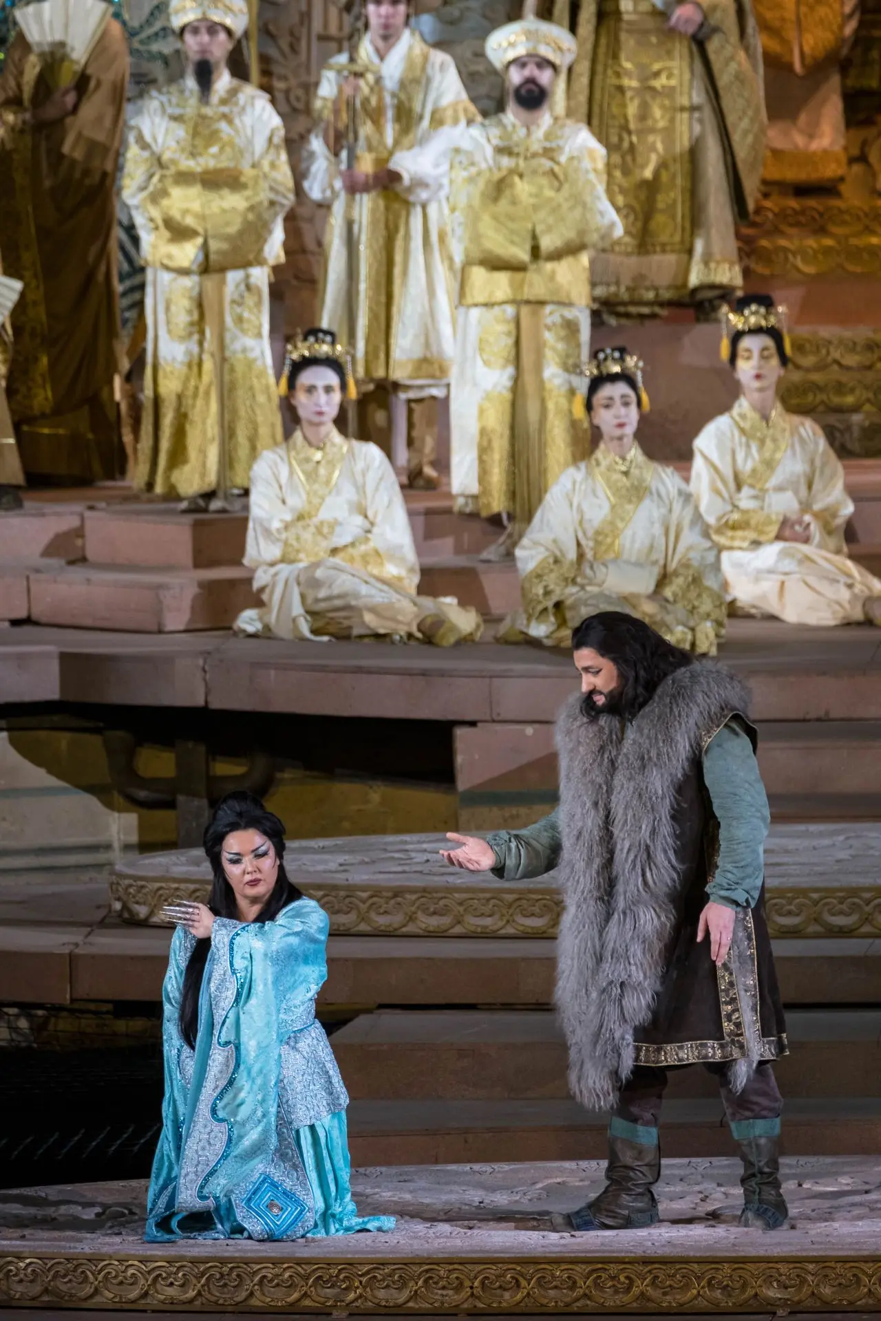 Al centro, in ginocchio, la principessa Turandot. Alla sua sinistra, Calaf, vestito come nell’immagine precedente. Turandot indossa una tunica celeste, glaciale, lunga fino ai piedi, con le maniche che si allargano all’altezza dei polsi e decorate con dei ricami ondulati di colore celeste, brillanti. Le spalline sono rinforzate, simili ad una corazza. La parte anteriore della tunica è decorata con delle fasce, anch’esse brillanti, sempre di colore celeste. Ai piedi, si intravedono solo le punte delle scarpe dello stesso colore della tunica. In testa, un copricapo a forma di raggiera, di colore argento. Le punte della raggera sono di colore blu. 