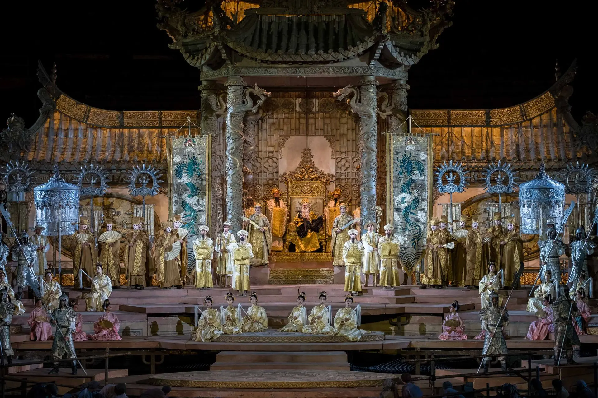 Il palcoscenico è occupato dalla reggia dell’imperatore Altoum, scintillante di oro e argento con riflessi verde giada. Il corpo centrale è costituito dal baldacchino del trono: una struttura che richiama il Baldacchino di San Pietro di Bernini, sostenuta da quattro colonne tortili, intorno a cui sono attorcigliati in spire dei draghi. Seduto sul trono, l’imperatore Altoum. A destra e a sinistra del baldacchino, gli stendardi dell’imperatore, uno per lato, di colore oro e argento, su cui è rappresentato un drago di colore blu. Di fronte, delle scalinate su cui si trova la folla: ballerine in rosa con ombrellini, comparse in bianco con pennacchi, dignitari in beige con ventagli, concubine dalle stoffe luccicanti. Questa scena crea un efficace contrasto tra lo spazio imperiale e quello popolare, che appare ancora più povero e spento.